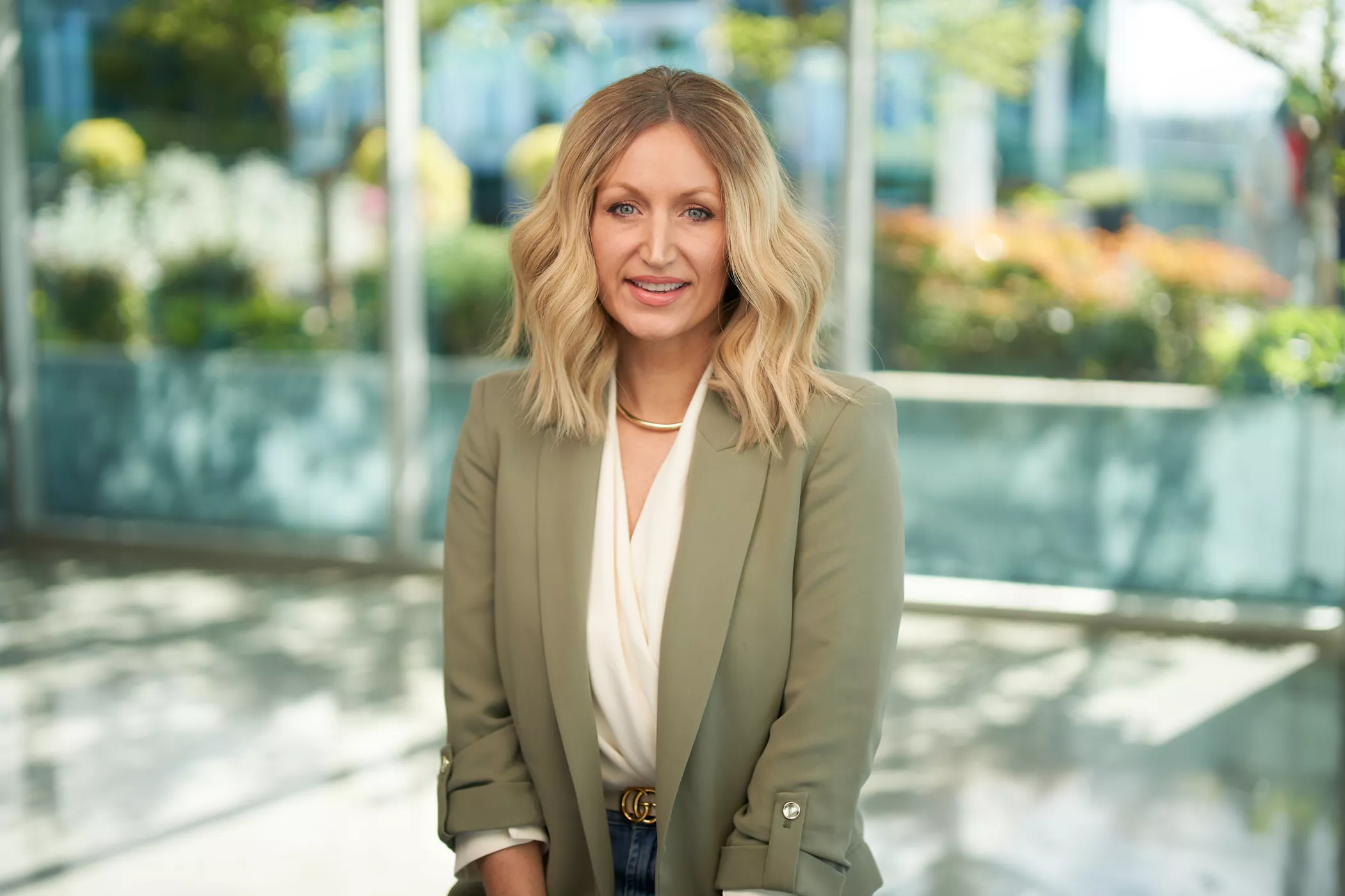 A woman wearing a green blazer and white blouse is seated in a modern indoor setting with large glass windows. The background features greenery and urban architecture, creating a bright and professional atmosphere. Her outfit includes a gold necklace and denim jeans, complementing the polished yet casual style.